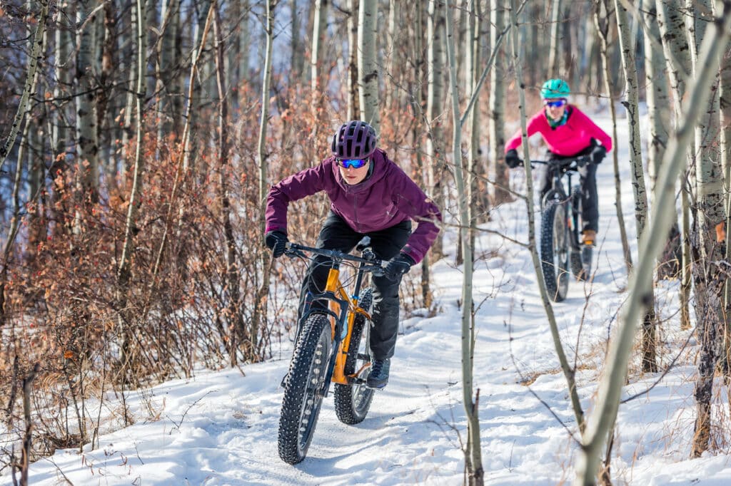Women using winter mountain bike tires to ride snowy trail.